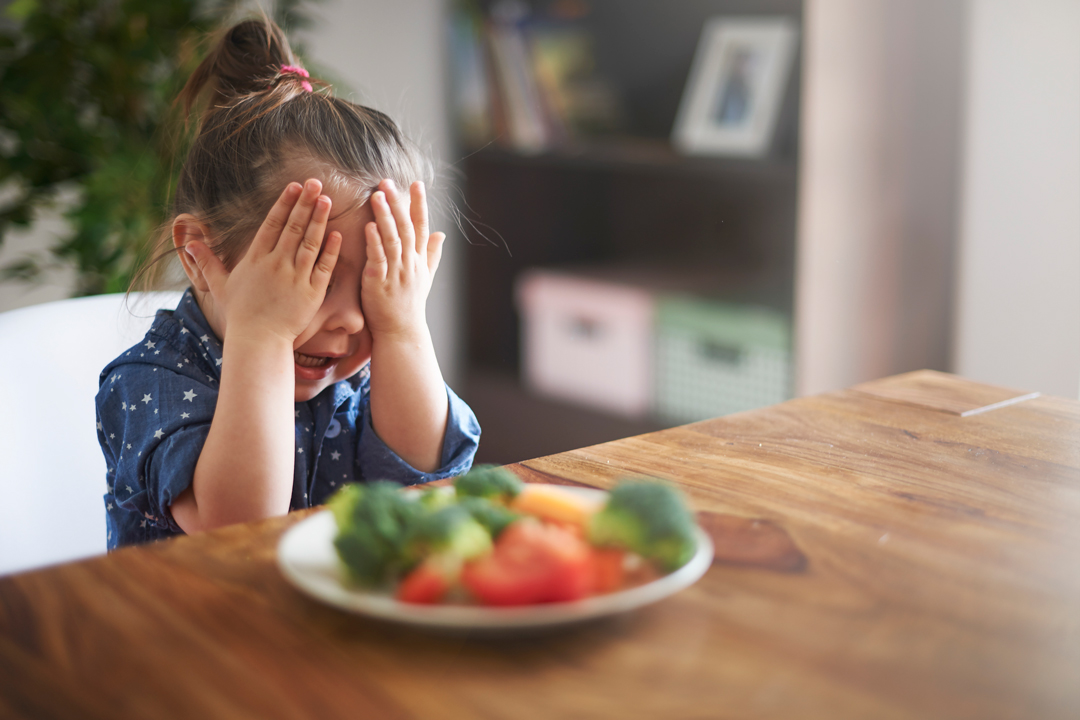 girl covering her eyes with a plate of vegetables in front